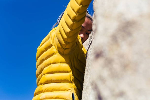 Petite donna asiatica arrampicata su roccia all'aperto pende da muro di pietra mentre indossa una giacca gialla gonfia brillante
 - Foto, immagini