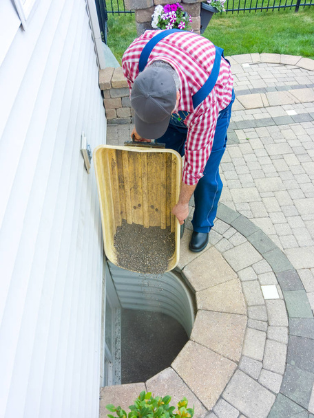 Man replacing the old pebbles in an egress window in the basement of his house during spring maintenance emptying the stones from a wheelbarrow - Photo, Image