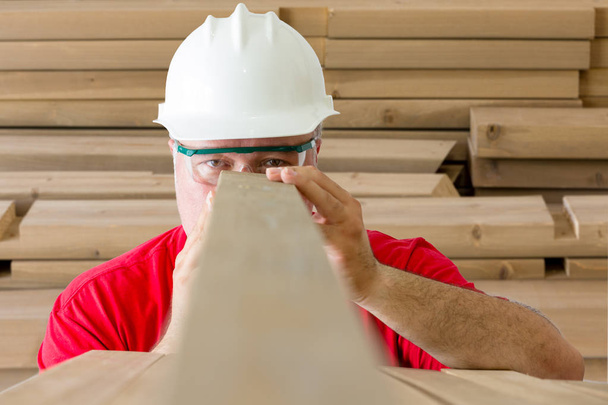 Uomo maturo con casco e occhiali di sicurezza che ispezionano la qualità della tavola di legno
 - Foto, immagini