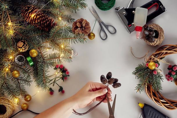 Vue de dessus des mains féminines font une couronne de Noël. Cadeaux et rouleaux emballés, branches d'épinette et outils sur table blanche
. - Photo, image