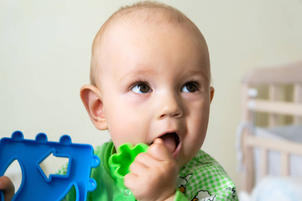 Ein einjähriges Baby nagt an einem Plastikspielzeug wegen seiner Kinderkrankheiten. kleiner fröhlicher Junge im hellgrünen Anzug mit Schafen. - Foto, Bild