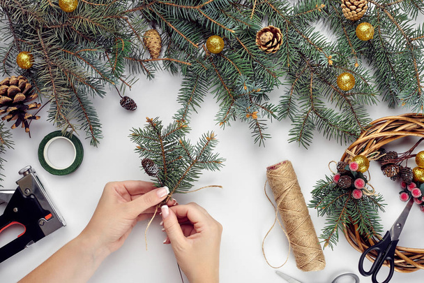 Vue de dessus des mains féminines font une couronne de Noël. Cadeaux et rouleaux emballés, branches d'épinette et outils sur table blanche
. - Photo, image