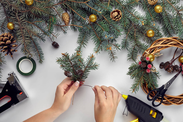 Верхний вид женских рук делает рождественский венок. Упакованные подарки и свитки, еловые ветви и инструменты на белом столе. Подготовка к празднику Рождества или Нового года
 - Фото, изображение