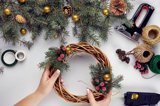 女性の手の平面図は、クリスマス リースを作る。ギフトやスクロールを満載、小ぎれいに枝と白いテーブル上のツール. - 写真・画像