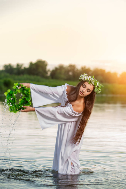 Menina de cabelos pretos bonita em vestido vintage branco e coroa de flores em pé na água do lago. Inflamação solar
. - Foto, Imagem