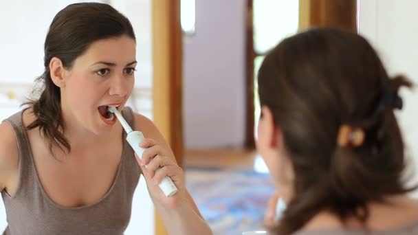 Young woman brushing teeth using electric toothbrush looking in the mirror - Filmagem, Vídeo
