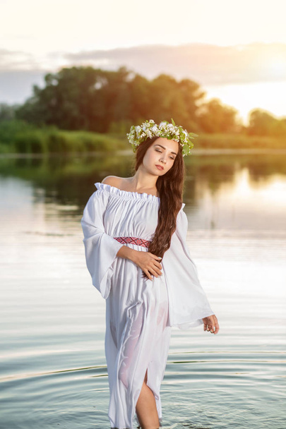 Beautiful black haired girl in white vintage dress and wreath of flowers standing in water of lake. Sun flare. - Photo, Image