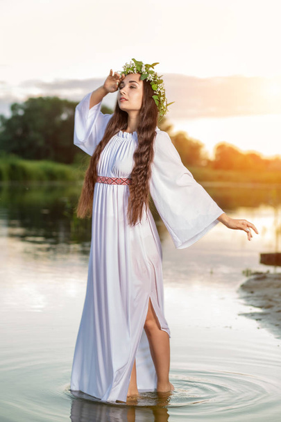 Belle fille aux cheveux noirs en robe vintage blanche et couronne de fleurs debout dans l'eau du lac. Éclair de soleil
. - Photo, image