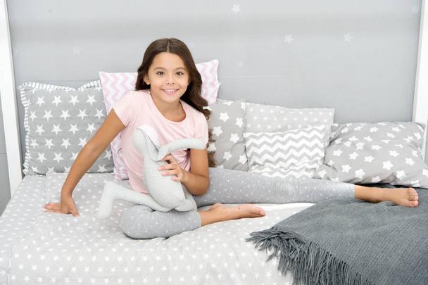 Girl long curly hair enjoy evening time with favorite toy. Kid sit bed and play bunny toy modern bedroom interior. Evening time. Girl child wear pajamas play bunny toy. Play soft toy before go sleep - Foto, Bild