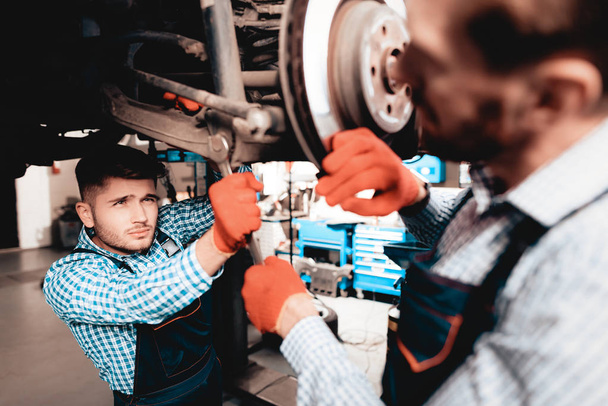 Young Mechanic Repairs Automotive Hub In Garage. Professional Uniform. Service Station Concept. Confident Engineer Stare. Detail Repairing. Under The Vehicle. Automobile Diagnostic. Join Forces. - 写真・画像