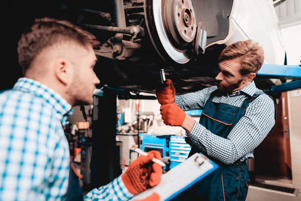 Young Mechanic Repairs Automotive Hub In Garage. Professional Uniform. Service Station Concept. Confident Engineer Stare. Detail Repairing. Under The Vehicle. Automobile Diagnostic. - Foto, Imagem