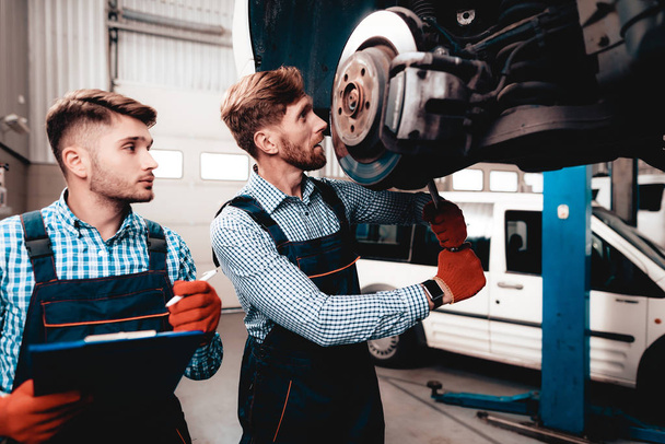 Young Mechanic Repairs Automotive Hub In Garage. Professional Uniform. Service Station Concept. Confident Engineer Stare. Detail Repairing. Under The Vehicle. Automobile Diagnostic. - Foto, immagini
