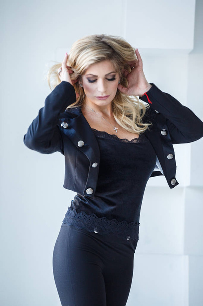 Fashion portrait of beautiful sexy blonde in black pants and black jacket standing on white studio background - 写真・画像