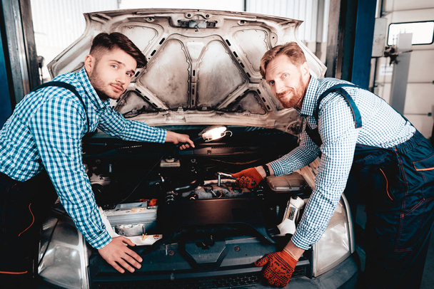 Young Auto Mechanics Repairs Car With A Wrench. Under The Hood. Professional Uniform. Using Flashlight. Engineering Specialist Team. Service Station. Automobile Diagnostic. Working In Garage. - Φωτογραφία, εικόνα