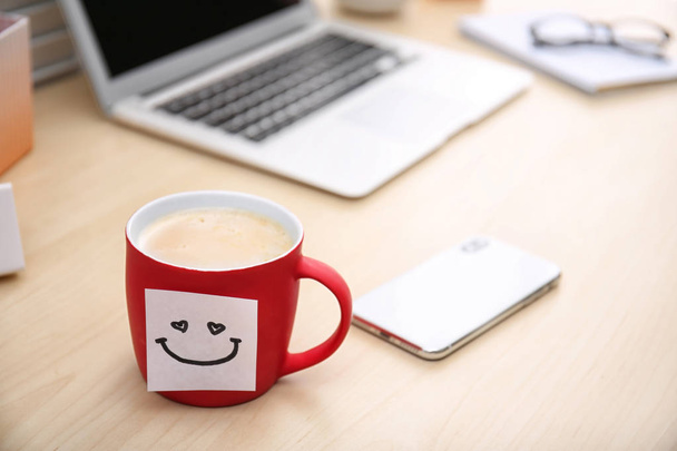 Sticky note with funny face attached to cup of coffee on office table. Space for text - Foto, Bild