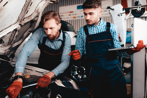 Two Auto Mechanics Work Together In Garage. Wrench Using. Laptop Screen. Under The Hood. Professional Uniform. Red Protective Gloves. Service Station Concept. Automobile Diagnostic. - Фото, изображение