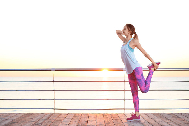 Young woman doing fitness exercises on pier in morning - Фото, изображение