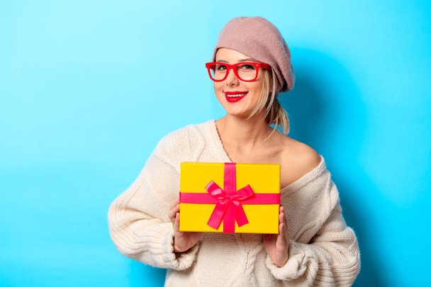 Portrait of a young girl in white sweater with yellow gift box on blue background - Photo, Image