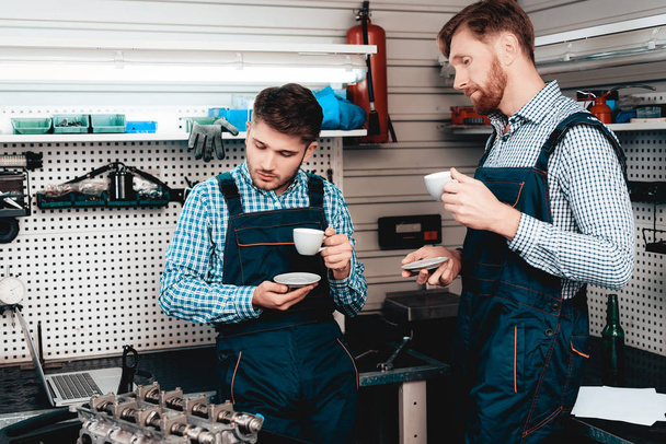 Two Young Cheerful Mechanics Are Drinking Coffee On A Service Station. Relaxing Concept. Having A Break. Cup And Saucer. Colleagues Discussion. Professional Uniform. Working In A Garage. - Фото, зображення