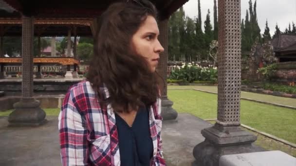 Aantrekkelijk meisje met krullend haar zit in arbor in balinese park op zomerdag - Video