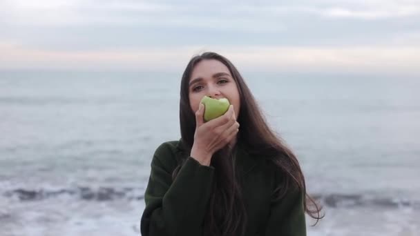 日の出ビーチで青リンゴを噛む暖かいコートで美しい少女のクローズ アップ - 映像、動画