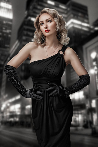 Das Retro-Porträt einer unerreichbaren schönen Frau im schwarzen Kleid mit roten Lippen, Smokey Eyes und langen Ohrringen steht vor dem Hintergrund der nächtlichen Stadt. film noir. vintage hollywood style. - Foto, Bild