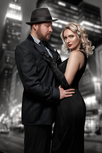 Film noir. Detective man in a raincoat and hat and a dangerous woman with red lips in black dress. Couple stands against the background of the night city - Фото, изображение