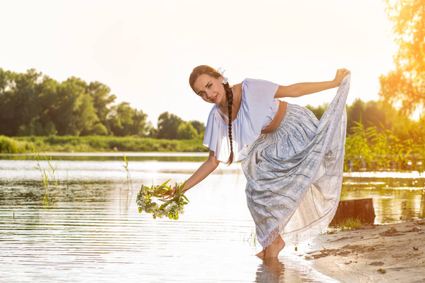 Young beautiful caucasian woman standing at the bank of river. Traditional countryside picture with girl at foreground and copy space. Sun flare - Photo, Image