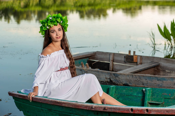 Young woman with flower wreath on her head, relaxing on boat on river at sunset. Concept of female beauty, rest in the village - Photo, Image