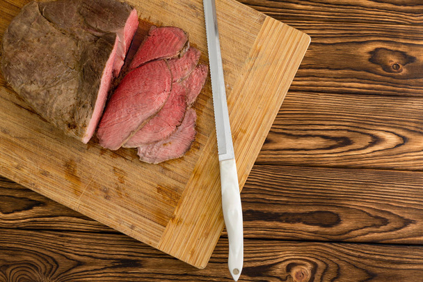 Overhead view of sliced meat sitting on top of cutting board next to knife over wooden grain background - Foto, Imagen