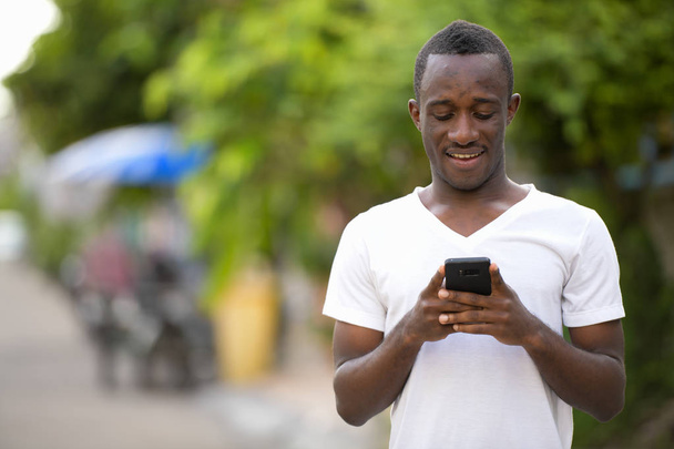 Молодой счастливый африканский мужчина улыбается, пользуясь телефоном на улице
 - Фото, изображение