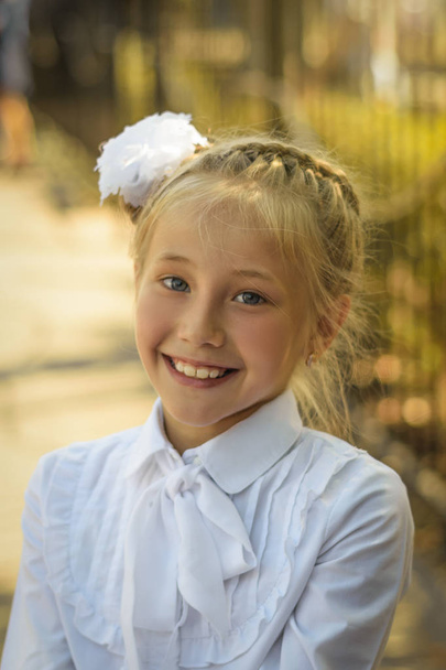 Una bella ragazza con un fiocco bianco sulla testa ride allegramente prima di andare a scuola. Ritratto di un bambino in età scolare che gioca nel parco autunnale
. - Foto, immagini