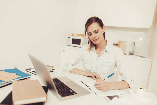 Young Woman Is Making Notes At The Kitchen. Work From Home With A Laptop Concept. Problems Solving. Official Clothes. Comfortable Environment. Calmness And Confidence. Concentrated Worker. - Photo, Image
