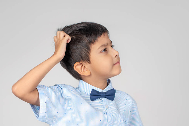 Le garçon portant une chemise bleue avec nœud papillon pense à quelque chose qui lui gratte la tête sur un fond gris dans le studio
 - Photo, image