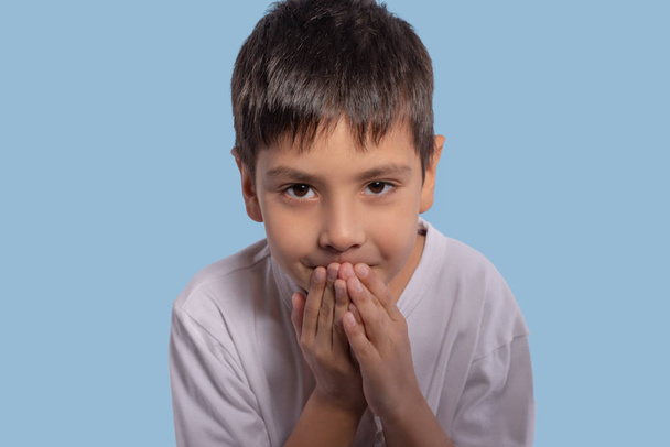 Nahaufnahme emotionales Porträt eines kleinen Jungen mit weißem Hemd auf blauem Hintergrund im Studio. er will ein Geheimnis teilen, also beugte er sich vor und bedeckte seinen Mund mit den Händen, um ein Geheimnis zu flüstern - Foto, Bild