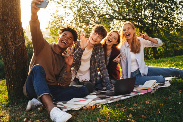 Group of cheerful multhiethnic students doing homework together at the park, using laptop computer, taking a selfie with mobile phone - Valokuva, kuva