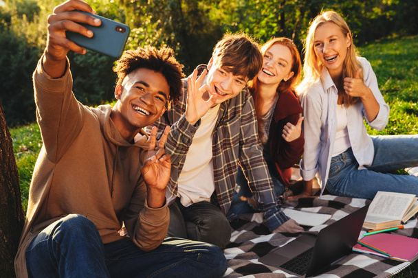 Group of positive multhiethnic students doing homework together at the park, using laptop computer, taking a selfie with mobile phone - Foto, Bild
