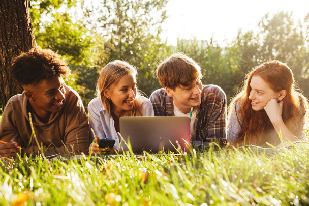 Group of excited multhiethnic students doing homework together at the park, using laptop computer, laying on a grass - Fotografie, Obrázek