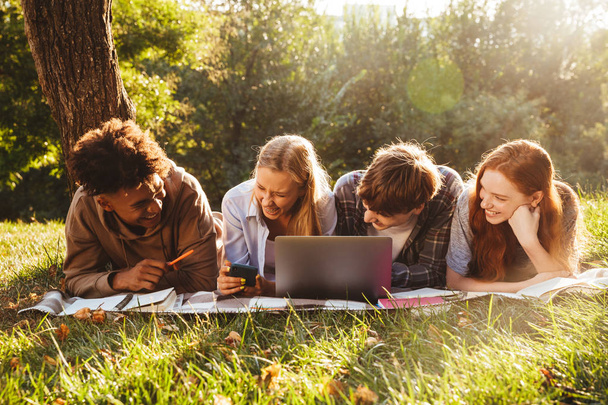 Group of laughing multhiethnic students doing homework together at the park, using laptop computer, laying on a grass - Photo, Image