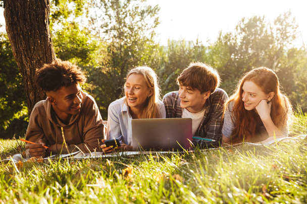 Group of joyful multhiethnic students doing homework together at the park, using laptop computer, laying on a grass - Fotografie, Obrázek