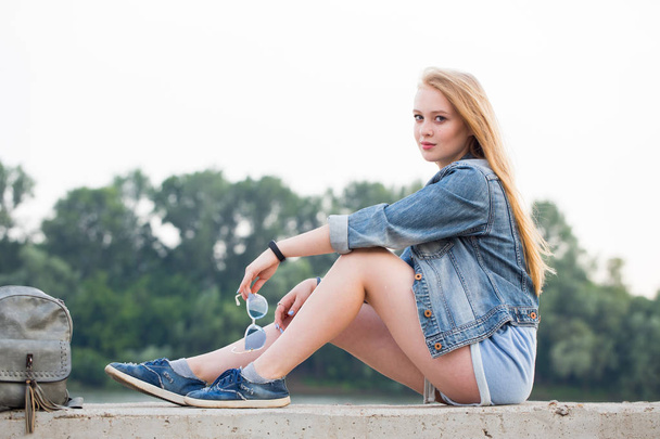 Young beautiful blonde woman in short shorts and jeans sitting on the waterfront in summer and looking at the camera. Student life - Foto, immagini