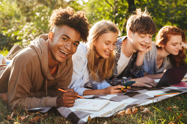 Group of cheerful multhiethnic students doing homework together at the park, using laptop computer and mobile phone, laying on a grass - Fotografie, Obrázek