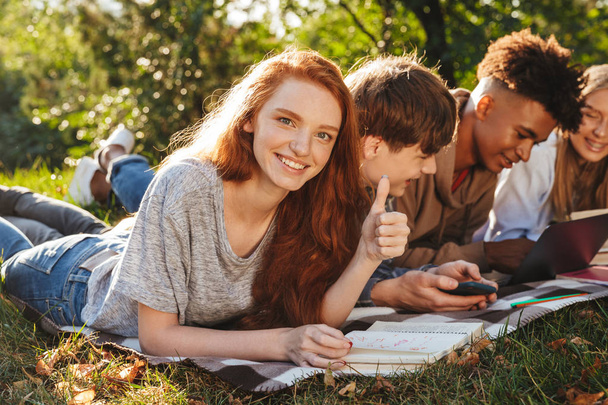 Group of energetic multhiethnic students doing homework together at the park, using laptop computer and mobile phone, laying on a grass - Fotografie, Obrázek
