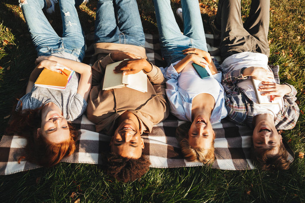 Vista superior del grupo de estudiantes multiétnicos felices tendidos en una hierba, sosteniendo libros
 - Foto, imagen