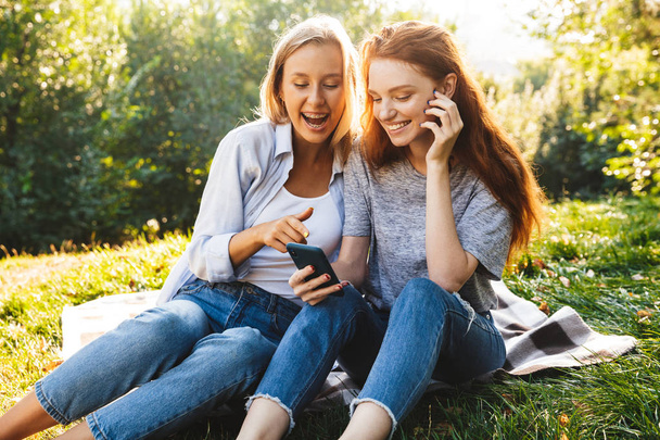 Image of pretty women students wearing casual clothes sitting on grass and using smartphone while resting in summer park - Foto, Bild