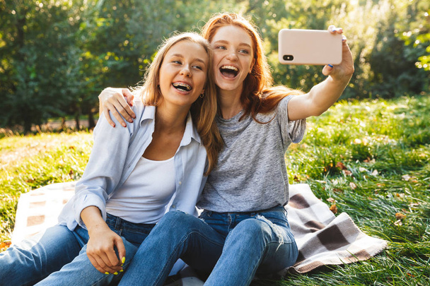 Image of two joyful women wearing casual clothes laughing, while sitting on grass and taking selfie on mobile phone while resting in summer park - Foto, immagini