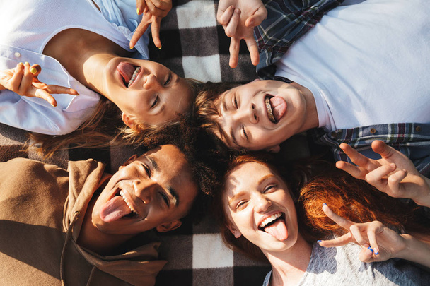 Imagen de cuatro personas alegres hombres y mujeres riendo y acostados en una manta en círculo al aire libre
 - Foto, Imagen