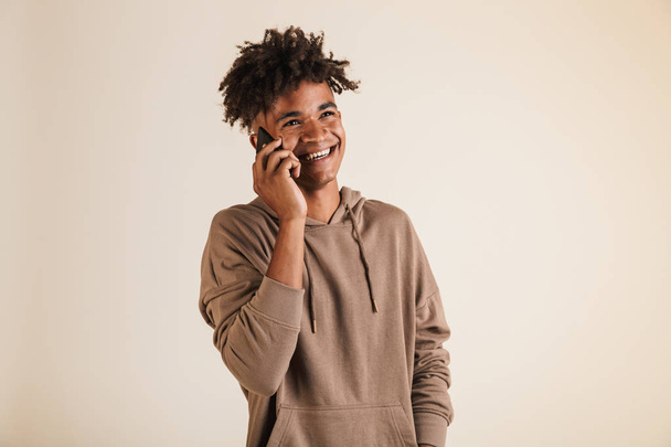 Portrait of a cheerful young afro american man dressed in hoodie isolated, talking on mobile phone - Fotografie, Obrázek
