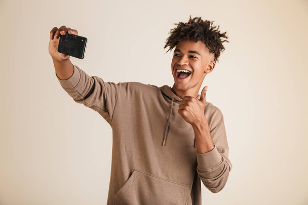 Foto eines fröhlichen jungen Afroamerikaners in Kapuzenpulli, der ein Selfie macht, während er Daumen nach oben zeigt. - Foto, Bild
