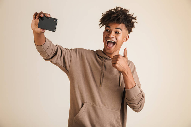 Photo of a cheerful young afro american man dressed in hoodie taking a selfie isolated while showing thumbs up gesture. - Foto, Bild
